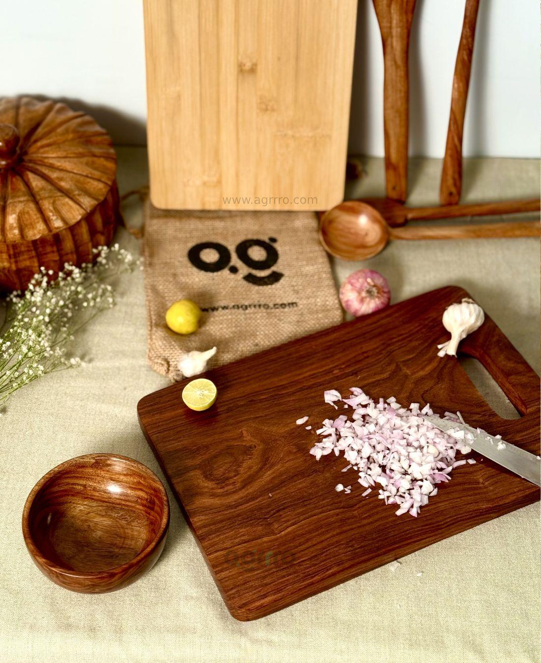 A rustic kitchen setup with a Sheesham wood chopping board featuring chopped onions and a kitchen knife, placed beside colorful vegetables and wooden kitchen utensils.