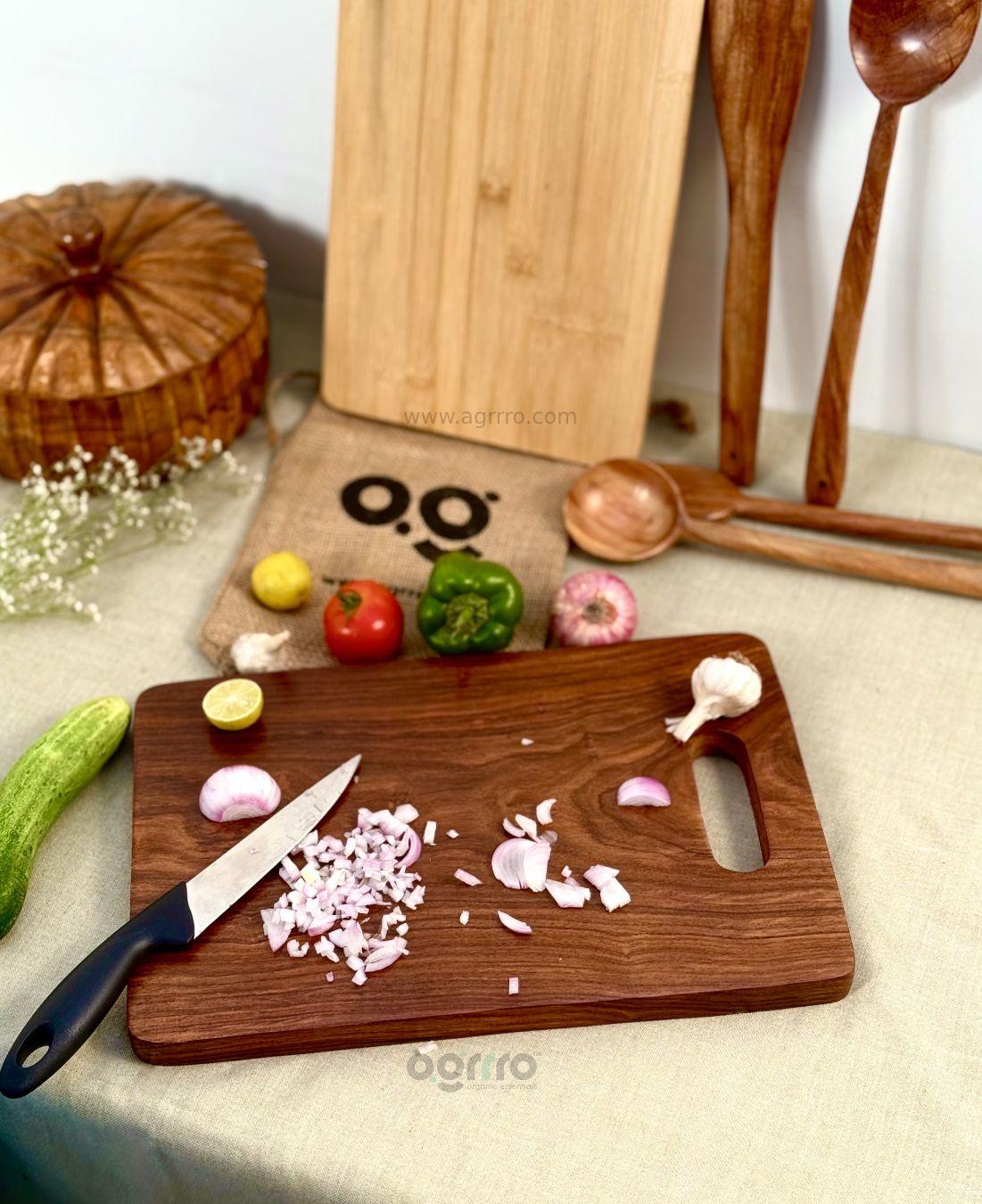 A rustic kitchen setup with a Sheesham wood chopping board featuring chopped onions and a kitchen knife, placed beside colorful vegetables and wooden kitchen utensils.