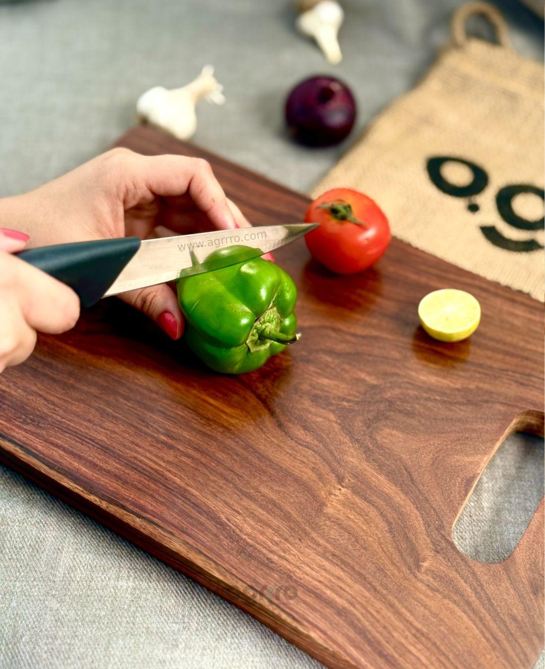 A close-up of a person slicing a green capsicum on a handcrafted Sheesham wood chopping board, surrounded by fresh vegetables like tomato, garlic, and lemon.